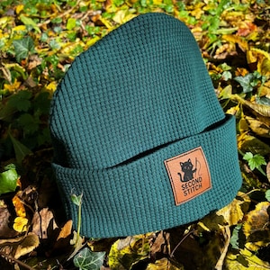Könnte beinhalten: Dunkelgrüne Beanie-Mütze mit strukturiertem Waffelstrick-Design. Die Mütze hat einen braunen Lederaufnäher mit einer schwarzen Katzen-Grafik und den Worten "SECOND STITCH". Die Mütze ist am Rand gefaltet.