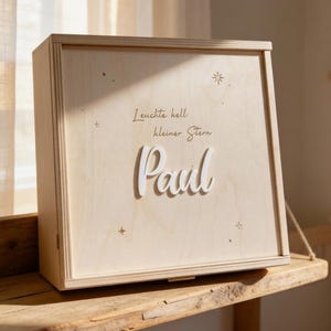 Könnte beinhalten: Eine hellfarbene Holzkiste mit dem Namen "Paul" in Schreibschrift. Die Kiste trägt den Text "Leuchte hell kleiner Stern" und Sterndesigns. Die Kiste steht auf einem Holzregal.