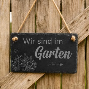 Peut inclure: Une plaque rectangulaire en ardoise noire suspendue par une corde en jute. La plaque porte l'inscription grav&eacute;e "Wir sind im Garten" dans un m&eacute;lange de polices, accompagn&eacute;e d'une illustration au trait de fleurs sauvages, d'un papillon et d'une abeille.