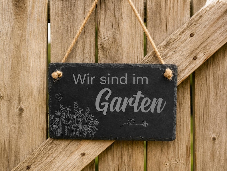 Peut inclure: Une plaque rectangulaire en ardoise noire suspendue par une corde en jute. La plaque porte l'inscription grav&eacute;e "Wir sind im Garten" dans un m&eacute;lange de polices, accompagn&eacute;e d'une illustration au trait de fleurs sauvages, d'un papillon et d'une abeille.