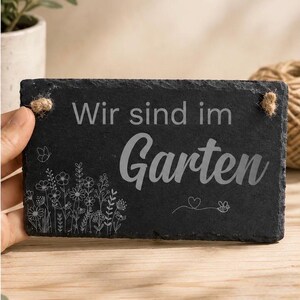 Peut inclure: Une plaque rectangulaire en ardoise noire aux bords irr&eacute;guliers. Le texte "Wir sind im Garten" est grav&eacute; au centre. Sous le texte, un dessin au trait blanc repr&eacute;sente un bouquet de fleurs sauvages, un petit c&oelig;ur et une abeille en vol. Deux cordes de jute sont fix&eacute;es aux coins sup&eacute;rieurs.