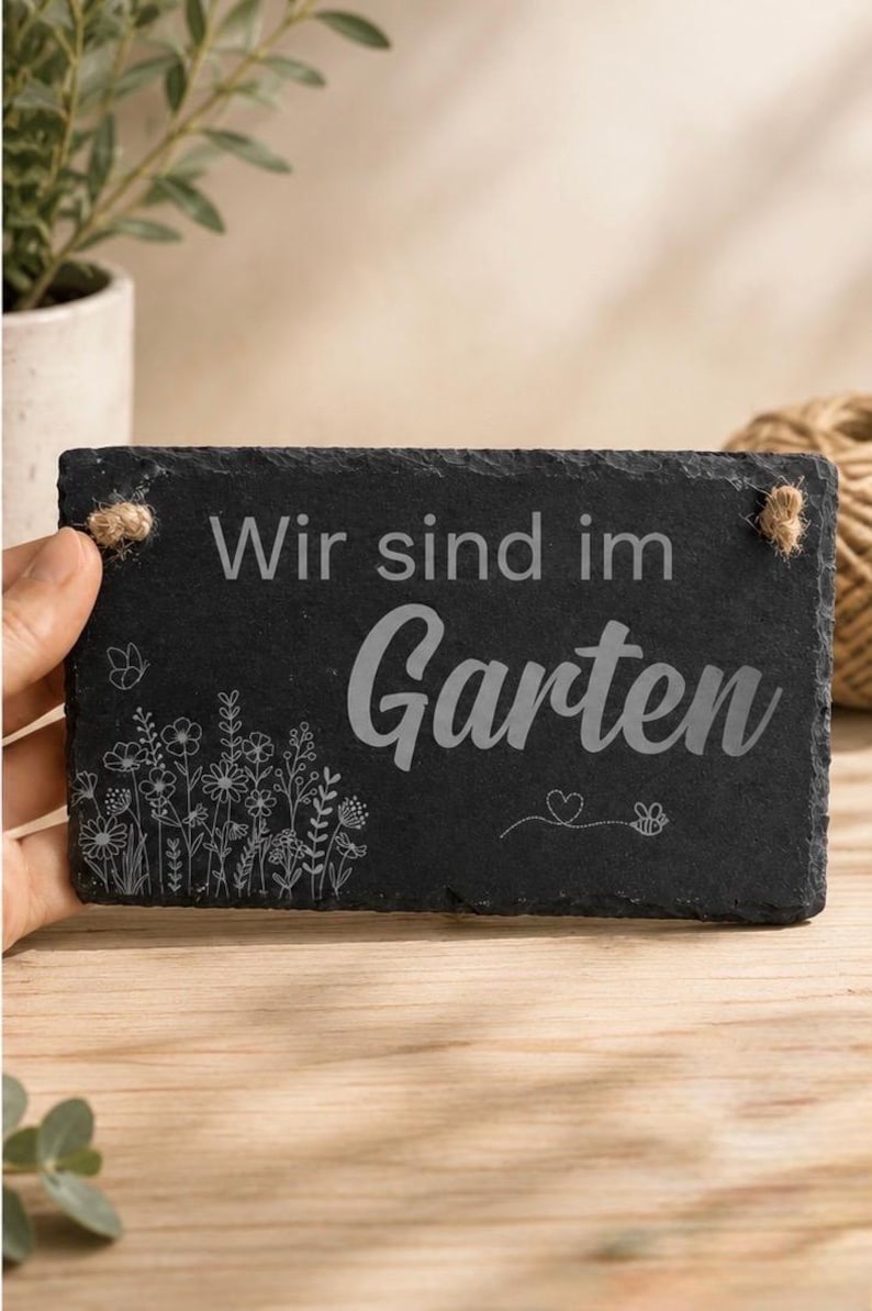Peut inclure: Une plaque rectangulaire en ardoise noire aux bords irr&eacute;guliers. Le texte "Wir sind im Garten" est grav&eacute; au centre. Sous le texte, un dessin au trait blanc repr&eacute;sente un bouquet de fleurs sauvages, un petit c&oelig;ur et une abeille en vol. Deux cordes de jute sont fix&eacute;es aux coins sup&eacute;rieurs.