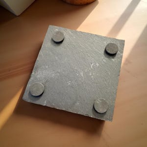 May include: A square, gray slate coaster with rounded corners. Four small, circular, gray pads are adhered to the underside. The coaster is designed to protect surfaces from heat and moisture.