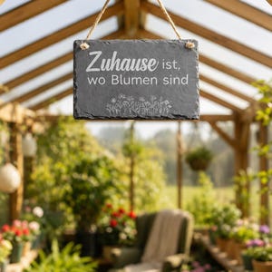 Peut inclure: Une plaque rectangulaire en ardoise gris fonc&eacute; suspendue par une corde en jute. La plaque pr&eacute;sente un texte blanc indiquant "Zuhause ist, wo Blumen sind" au-dessus d'une illustration au trait blanc de fleurs sauvages et d'un arrosoir.