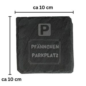 May include: A square, dark gray slate coaster, approximately 4 inches in size, with the letter "P" and the words "PF&Auml;NNCHEN PARKPLATZ" etched into the surface. The coaster has a rough, natural edge.