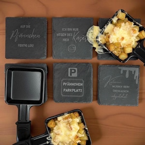 May include: A tabletop raclette grill with two filled pans of melted cheese and potatoes. The image also shows six slate coasters with engraved German text, including phrases like "Pf&auml;nnchen Parkplatz" and "Ich bin nur wegen des K&auml;ses hier."