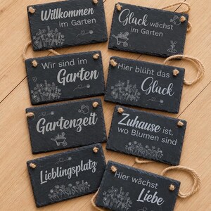 Peut inclure: Huit plaques rectangulaires en ardoise aux bords irr&eacute;guliers, chacune orn&eacute;e de textes allemands grav&eacute;s en blanc et d'illustrations botaniques. Les plaques portent des phrases comme Willkommen im Garten et Liebe, fix&eacute;es par des cordes en jute.