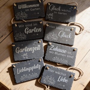 Peut inclure: Un ensemble de huit plaques rectangulaires en ardoise aux bords irr&eacute;guliers avec des cordes de suspension en jute. Chaque pierre gris fonc&eacute; pr&eacute;sente des gravures blanches avec du texte et des illustrations botaniques, incluant les phrases Willkommen im Garten, Gl&uuml;ck w&auml;chst im Garten, Wir sind im Garten, Hier bl&uuml;ht das Gl&uuml;ck, Gartenzeit, Zuhause ist wo Blumen sind, Lieblingsplatz et Hier w&auml;chst Liebe.