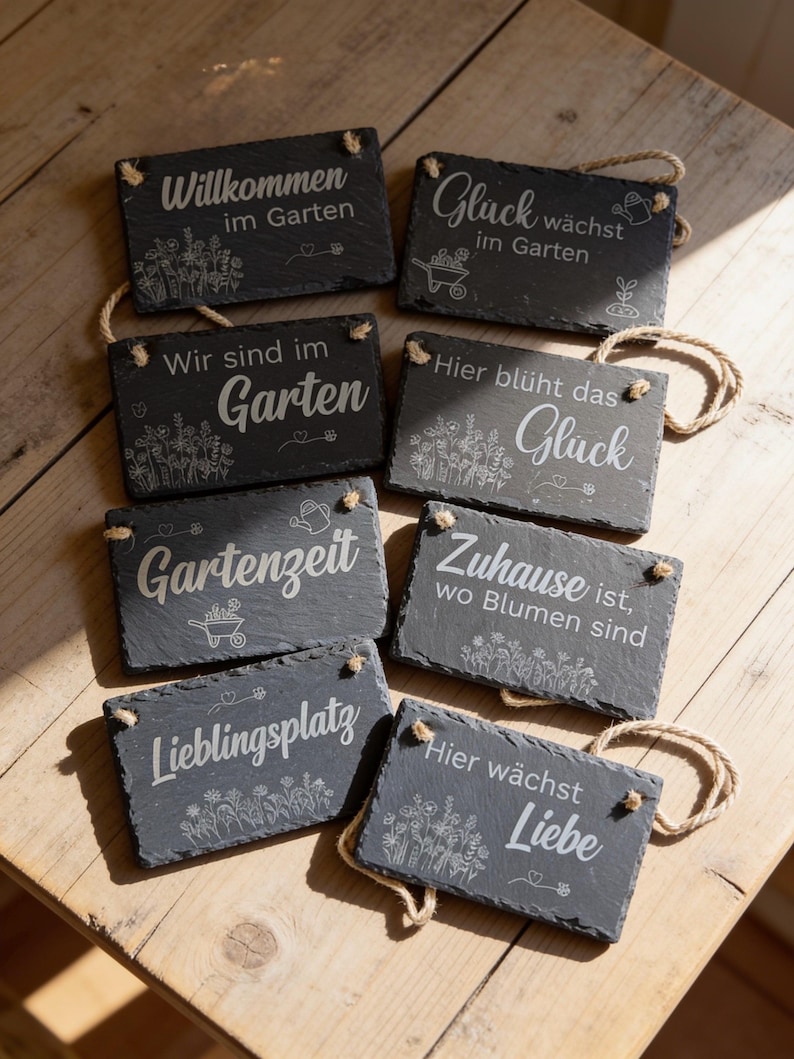 Peut inclure: Un ensemble de huit plaques rectangulaires en ardoise aux bords irr&eacute;guliers avec des cordes de suspension en jute. Chaque pierre gris fonc&eacute; pr&eacute;sente des gravures blanches avec du texte et des illustrations botaniques, incluant les phrases Willkommen im Garten, Gl&uuml;ck w&auml;chst im Garten, Wir sind im Garten, Hier bl&uuml;ht das Gl&uuml;ck, Gartenzeit, Zuhause ist wo Blumen sind, Lieblingsplatz et Hier w&auml;chst Liebe.
