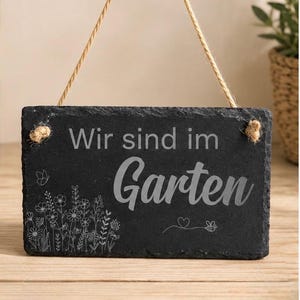 Peut inclure: Une plaque rectangulaire en ardoise noire aux bords irr&eacute;guliers, suspendue par une cordelette en jute beige. La plaque porte l'inscription grav&eacute;e 'Wir sind im Garten' en polices vari&eacute;es, accompagn&eacute;e de motifs floraux et d'une abeille.