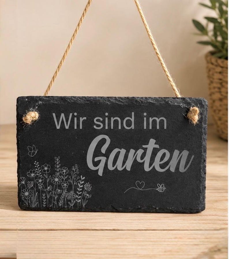 Peut inclure: Une plaque rectangulaire en ardoise noire aux bords irr&eacute;guliers, suspendue par une cordelette en jute beige. La plaque porte l'inscription grav&eacute;e 'Wir sind im Garten' en polices vari&eacute;es, accompagn&eacute;e de motifs floraux et d'une abeille.