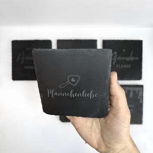 May include: A set of square, dark gray slate coasters. One coaster is held in a hand, displaying the text "Pf&auml;nnchenliebe" with a magnifying glass icon. Other coasters in the background have etched text. The coasters have a rough, textured surface.