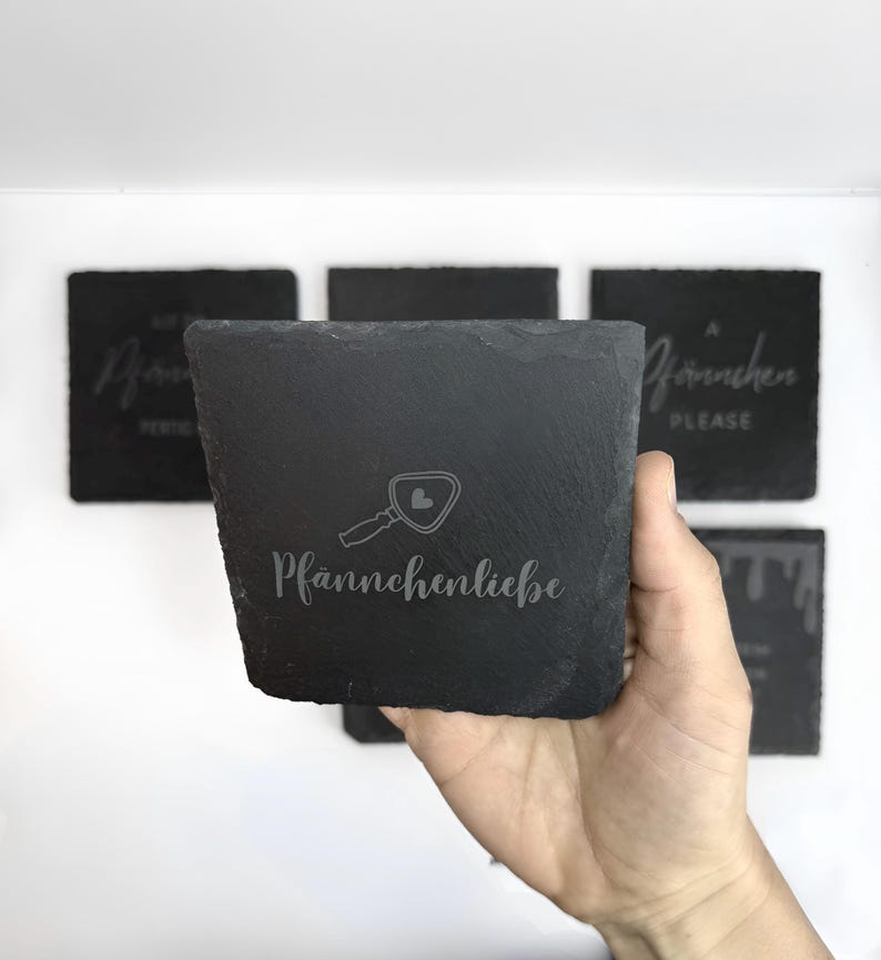 May include: A set of square, dark gray slate coasters. One coaster is held in a hand, displaying the text "Pf&auml;nnchenliebe" with a magnifying glass icon. Other coasters in the background have etched text. The coasters have a rough, textured surface.