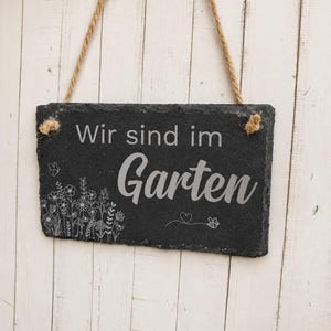 Peut inclure: Une plaque rectangulaire en ardoise noire suspendue par une corde en jute. La plaque pr&eacute;sente le texte grav&eacute; en blanc "Wir sind im Garten" accompagn&eacute; d'un dessin au trait d&eacute;taill&eacute; de fleurs sauvages, d'un papillon, d'un c&oelig;ur et d'une abeille.