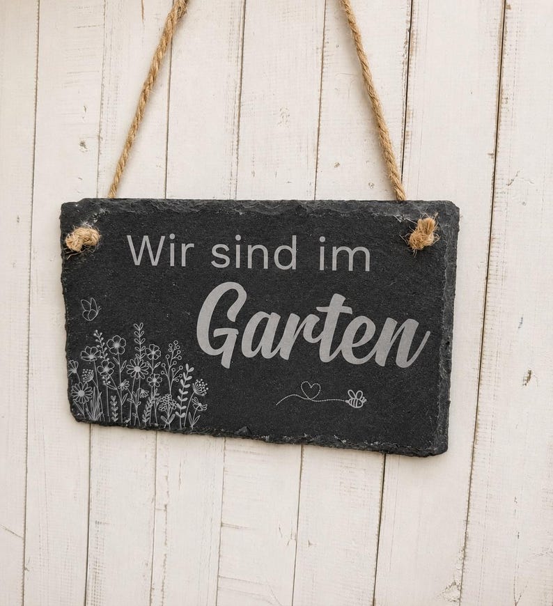Peut inclure: Une plaque rectangulaire en ardoise noire suspendue par une corde en jute. La plaque pr&eacute;sente le texte grav&eacute; en blanc "Wir sind im Garten" accompagn&eacute; d'un dessin au trait d&eacute;taill&eacute; de fleurs sauvages, d'un papillon, d'un c&oelig;ur et d'une abeille.