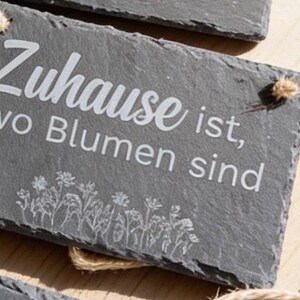 Peut inclure: Une plaque rectangulaire en ardoise gris fonc&eacute; avec le texte grav&eacute; en blanc "Zuhause ist, wo Blumen sind" et un dessin au trait de fleurs sauvages en bas. La plaque pr&eacute;sente un bord naturel brut et inclut une petite ficelle de jute.
