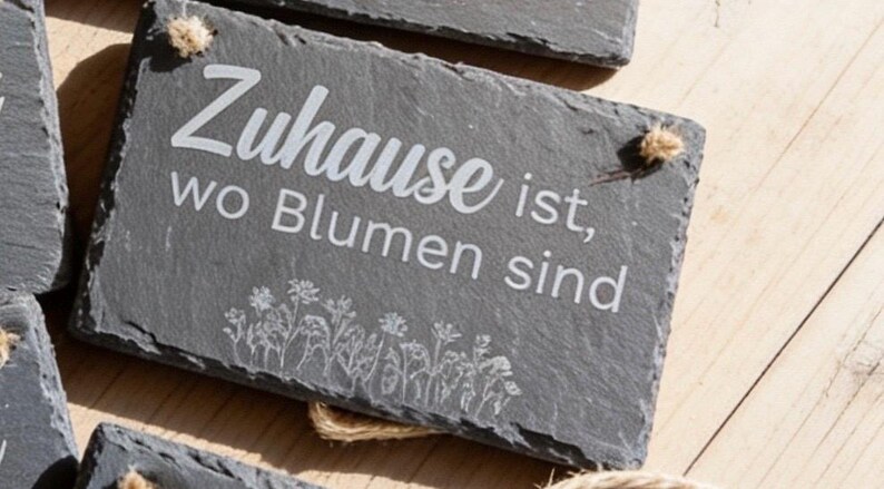 Peut inclure: Une plaque rectangulaire en ardoise gris fonc&eacute; avec le texte grav&eacute; en blanc "Zuhause ist, wo Blumen sind" et un dessin au trait de fleurs sauvages en bas. La plaque pr&eacute;sente un bord naturel brut et inclut une petite ficelle de jute.