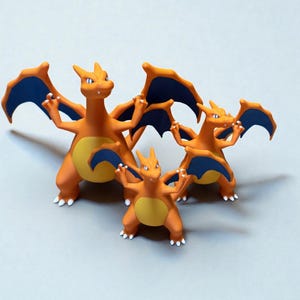 Könnte beinhalten: Drei orange und gelbe Charizard-Figuren mit blauen Flügeln werden vor einem hellgrauen Hintergrund gezeigt. Die Figuren sind in verschiedenen Größen, wobei die größte hinten und die kleinste vorne ist.