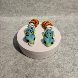Peut inclure: Une paire de boucles d'oreilles faites à la main en forme de gecko. Les boucles d'oreilles ont un corps bleu avec des taches noires, une queue tachetée blanche et orange et une boucle verte. Elles sont présentées sur un support circulaire blanc.