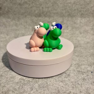 May include: A collection of colorful, cartoonish dinosaur figurines in pink, green, and blue. Each dinosaur has large white eyes and small feet. The figurines are displayed on a white, circular platform.