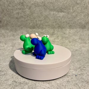 May include: Three colorful frog figurines. Two green frogs and one blue frog are positioned on a white, round platform. The frogs have white eyes and are set against a gray background. The figurines are whimsical and playful.