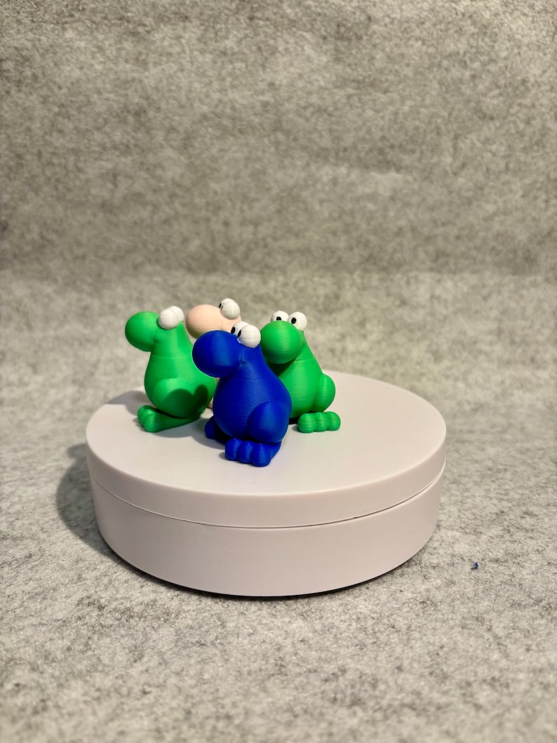 May include: Three colorful frog figurines. Two green frogs and one blue frog are positioned on a white, round platform. The frogs have white eyes and are set against a gray background. The figurines are whimsical and playful.
