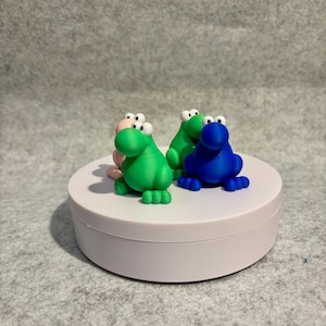 May include: Four colorful frog figurines in pink, green, and blue with large, googly eyes are displayed on a white, round platform. The frogs have detailed features and are arranged in a playful composition.