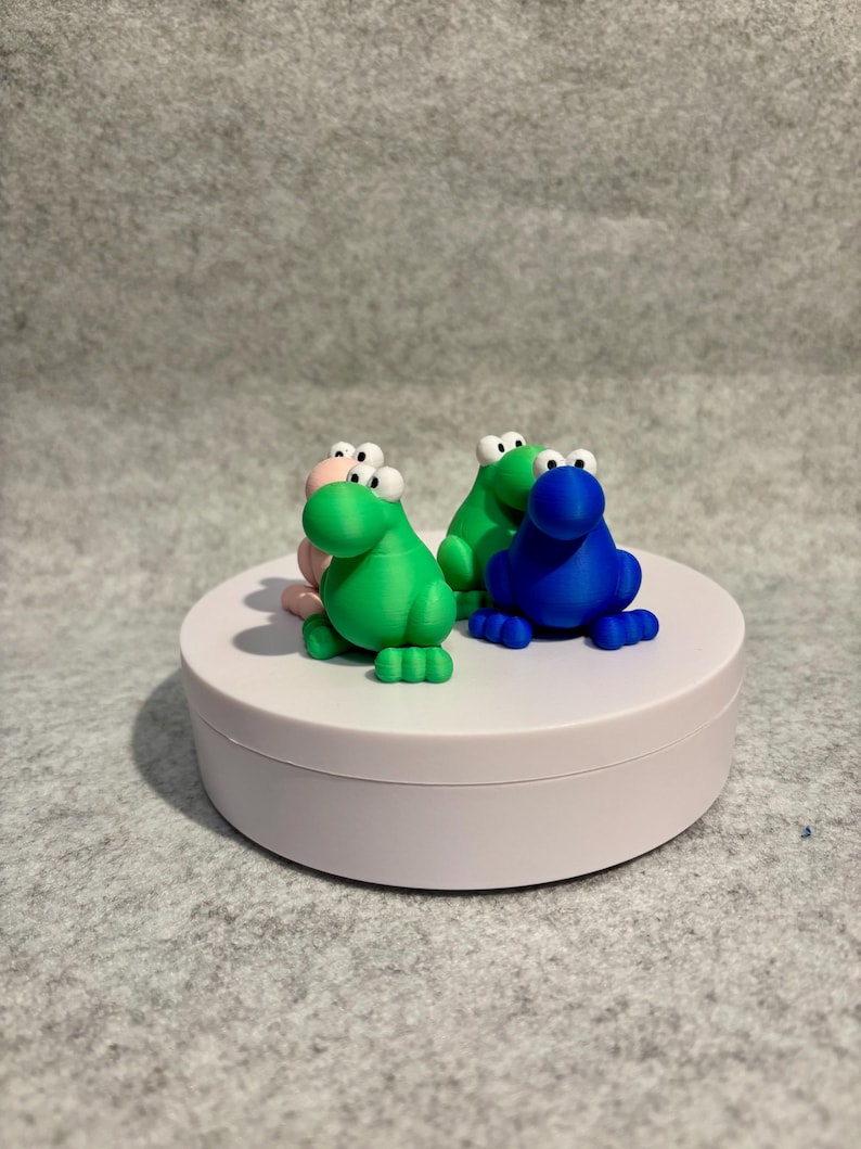 May include: Four colorful frog figurines in pink, green, and blue with large, googly eyes are displayed on a white, round platform. The frogs have detailed features and are arranged in a playful composition.