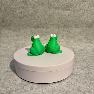 May include: Two bright green frog figurines with white eyes are displayed on a white circular platform. The frogs are facing away from the viewer, showcasing their backs. The background is a neutral gray.