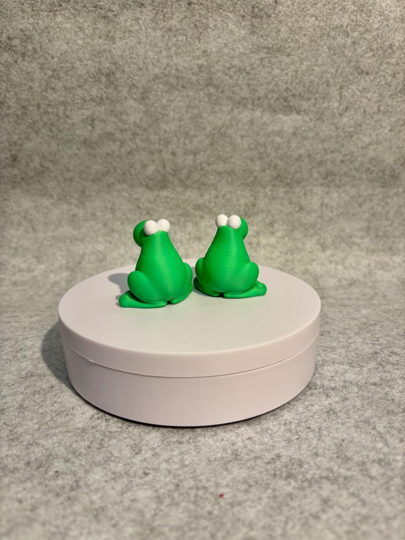 May include: Two bright green frog figurines with white eyes are displayed on a white circular platform. The frogs are facing away from the viewer, showcasing their backs. The background is a neutral gray.