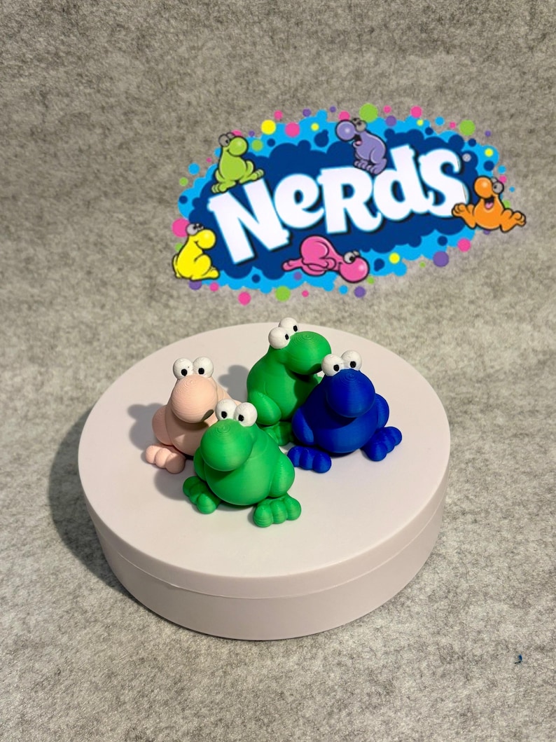 May include: Four colorful frog figurines in pink, green, and blue sit on a white platform. The frogs have large eyes and are positioned in a playful arrangement. A blue and white "Nerds" logo is in the background.