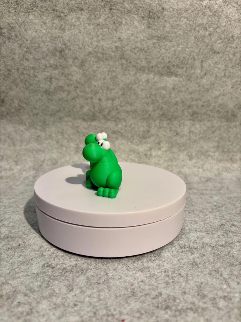 May include: A bright green frog figurine with white eyes and small white accents. The frog is sitting on a white, round platform. The background is a light gray fabric.
