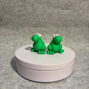 May include: Two bright green frog figurines with large white eyes and small feet are displayed on a light pink, round platform. The frogs are side-by-side, and the platform sits against a gray background.