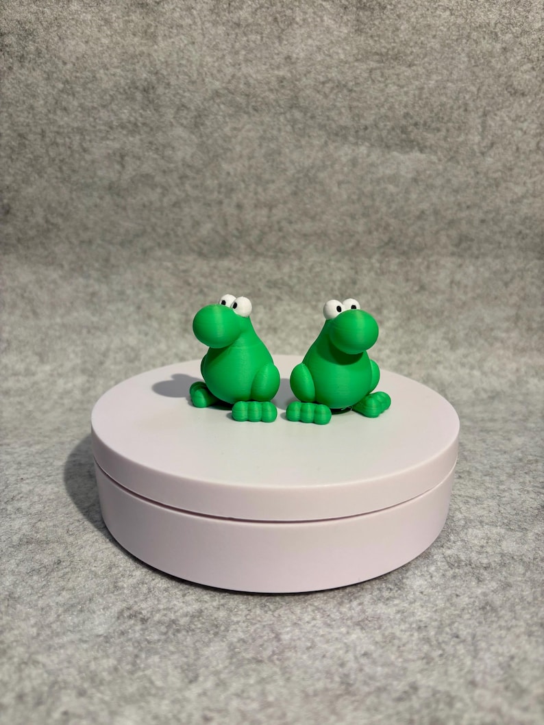 May include: Two bright green frog figurines with large white eyes and small feet are displayed on a light pink, round platform. The frogs are side-by-side, and the platform sits against a gray background.