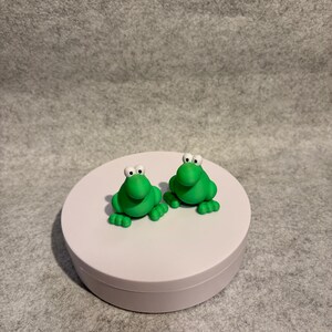 May include: Two small, bright green frog figurines with white eyes and black pupils sit side-by-side on a white, round platform. The frogs have four small feet and are made of a smooth material. The background is a neutral gray.