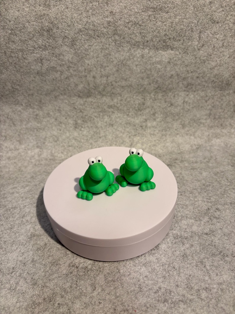 May include: Two small, bright green frog figurines with white eyes and black pupils sit side-by-side on a white, round platform. The frogs have four small feet and are made of a smooth material. The background is a neutral gray.