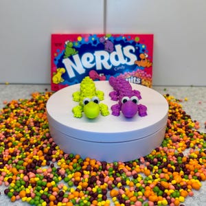 May include: A vibrant image showcases two small, 3D-printed alligator figurines in lime green and purple. They sit atop a white platform surrounded by colorful Nerds candy. A box of Nerds candy is in the background.