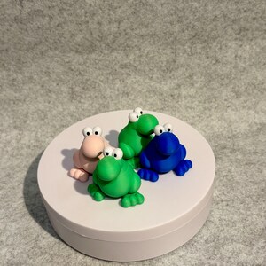 May include: Four colorful frog figurines in pink, green, and blue sit on a white platform. Each frog has large, white-eyed eyes and is posed in a sitting position. The background is a neutral gray.