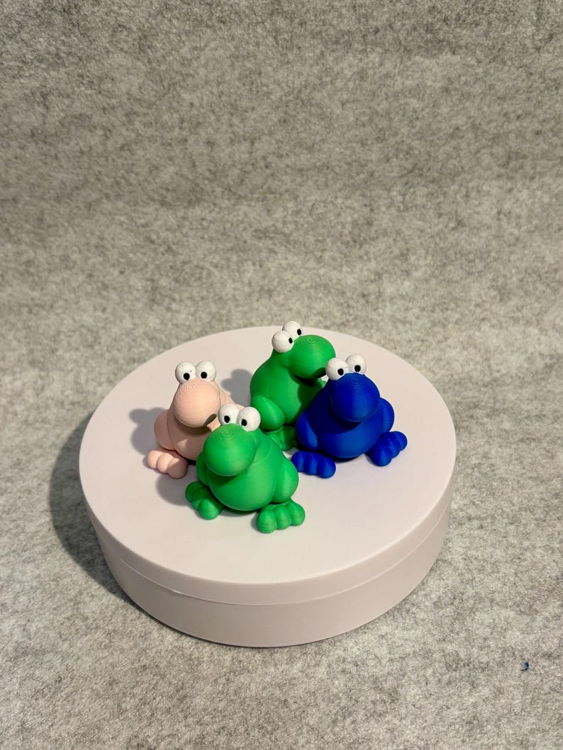 May include: Four colorful frog figurines in pink, green, and blue sit on a white platform. Each frog has large, white-eyed eyes and is posed in a sitting position. The background is a neutral gray.