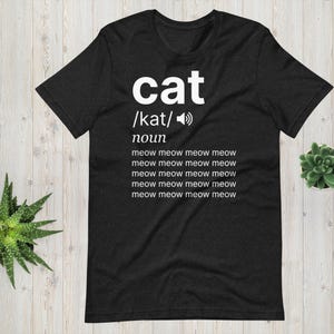 Peut inclure: T-shirt noir avec le mot "cat" en blanc, sa prononciation phonétique et le mot "nom". Le t-shirt porte le texte "meow meow meow meow" répété cinq fois. Le t-shirt est en matière souple.
