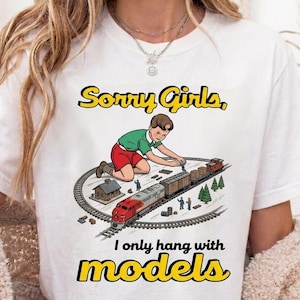 May include: White t-shirt with a graphic of a boy playing with a model train set. The text says "Sorry Girls, I only hang with models" in yellow and black. The design includes a train, tracks, and miniature buildings.