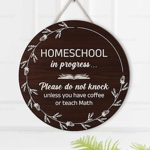 May include: Round, dark brown wooden sign with the text "HOMESCHOOL in progress... Please do not knock unless you have coffee or teach Math." The sign is decorated with a white floral design and hangs from a white door.