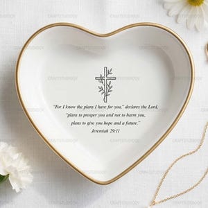 May include: A heart-shaped white ceramic trinket dish with a gold rim, featuring a cross design and a Bible verse. A gold ring and necklace with a pearl pendant are also present, along with white flowers.