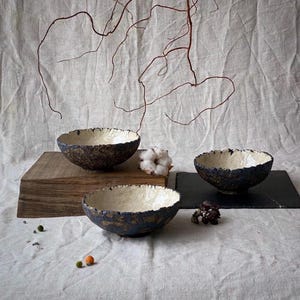 以下が含まれることがあります： 手作りのセラミックボウル3つ。外側は質感のある濃い青色、内側はオフホワイト。ニュートラルなリネンの上に配置され、木製のブロックや枝などの装飾要素が添えられています。