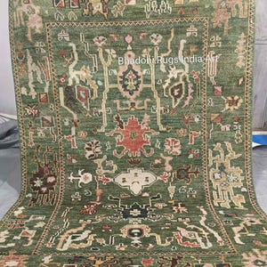 May include: A green area rug with an intricate, symmetrical pattern. The rug features a central medallion design with floral and geometric motifs in shades of red, brown, and cream. The text "Bhadohi Rugs India Art" is visible on the rug.
