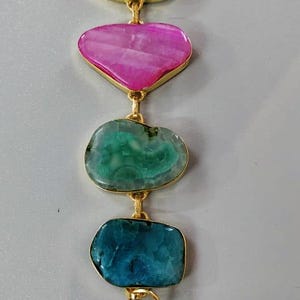 Könnte beinhalten: Goldfarbenes Armband mit fünf unregelmäßig geformten Steinen in Grün-, Rosa- und Blautönen. Die Steine sind in goldfarbenem Metall gefasst. Das Armband hat einen Verschluss.
