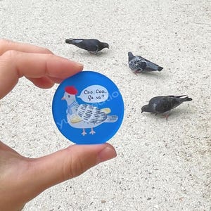 Peut inclure: Un badge rond bleu illustré d'un pigeon gris et blanc portant un béret rouge et une écharpe rayée bleue et blanche. Le pigeon est représenté avec une bulle de dialogue contenant le texte suivant : Coo... Coo... Ça va?