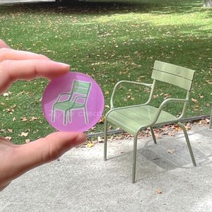 Puede incluir: Un disco circular de acrílico morado translúcido con un gráfico de contorno blanco de una silla de metal verde. El disco muestra el texto Zilye creation en letras blancas en el centro, posicionado frente a una silla verde a juego.