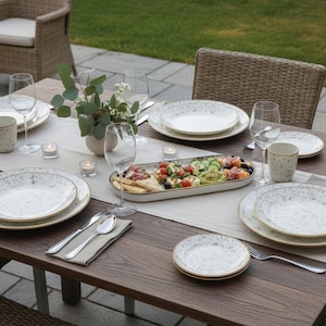 Puede incluir: Una mesa de madera preparada para una comida al aire libre. La mesa está puesta con platos, vasos, cubiertos y una fuente de comida. Los platos tienen un diseño moteado con un borde dorado. Un jarrón con vegetación y velas también están sobre la mesa.