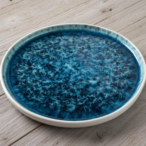 Puede incluir: Un plato redondo de cerámica con un esmalte azul profundo texturizado. El plato tiene un borde blanco y un patrón moteado en tonos azules y blancos. El plato está sobre una superficie de madera.